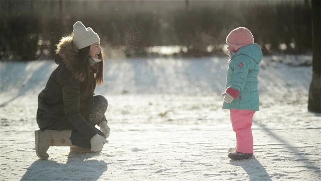 Laughing Woman in Warm Clothing is Throwing Snow at Her Daughter Wearing Snowsuit. Mother and Child Enjoying Cold Sunny Winter Morning Outdoors.