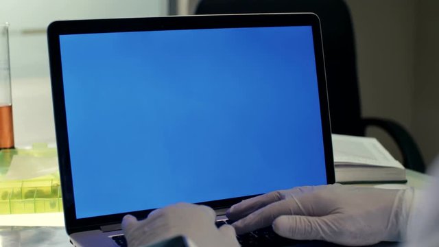 Elderly doctor working on laptop with blue screen in modern office on a background of test tubes