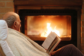 Happy senior man, wrapped in warm knitted plaid, relaxing at home in the evening, sitting in rocking chair by fireplace and reading a book - successful retirement concept