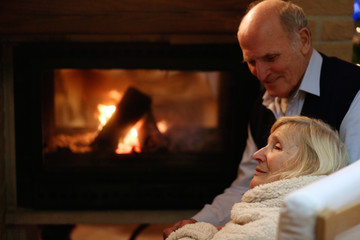 Happy senior couple, wrapped in warm knitted plaid, relaxing at home in the living room, sitting in rocking chair by fireplace and enjoying cosy evening together - successful retirement concept