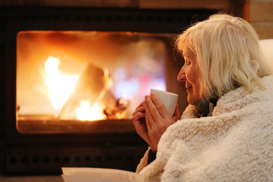Happy senior woman, wrapped in warm knitted plaid, relaxing at home in the evening, sitting in rocking chair, drinking tea, reading book and enjoying fireplace - successful retirement concept