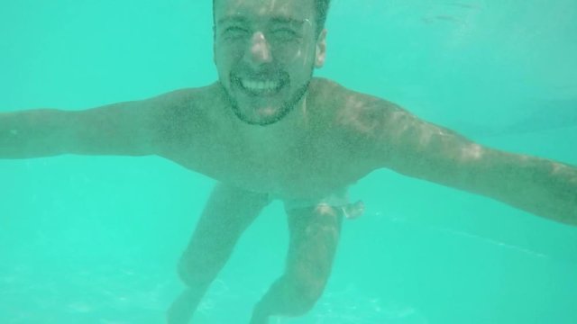Funny Caucasian topless male with light dark beard diving in pool smiling laughing. Close up underwater footage in slowmotion