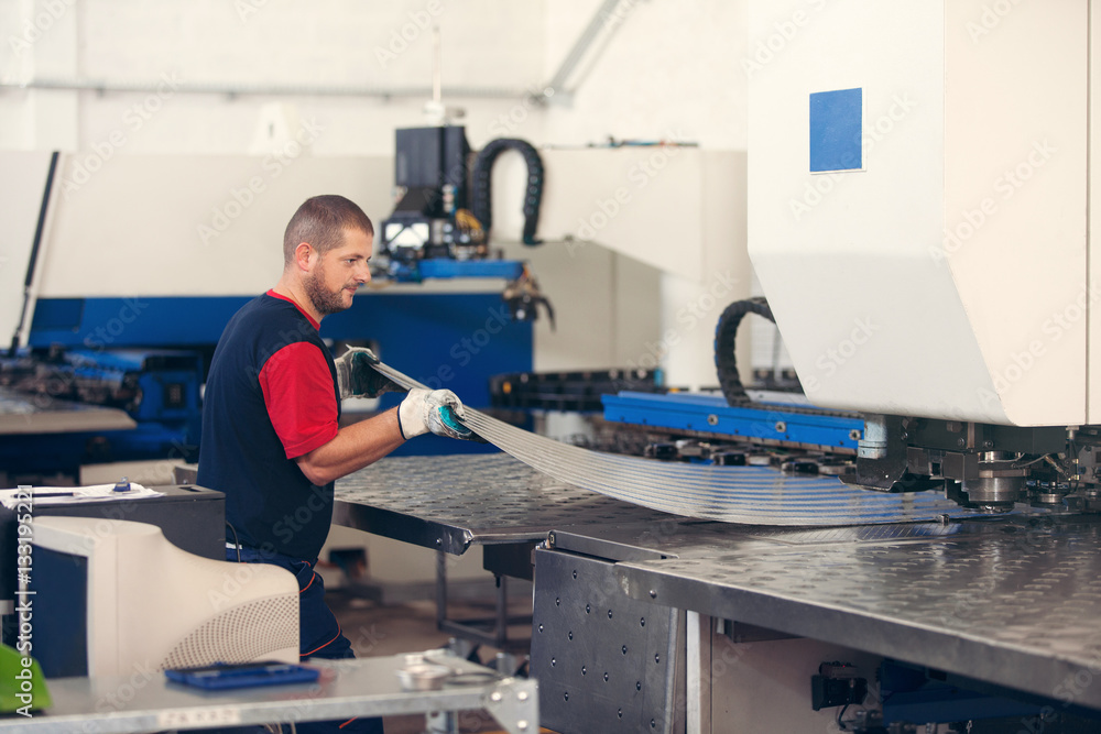 Wall mural inside a factory, industrial worker in action on metal press machine holding a piece of steel ready 