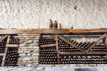 Old wine cellar with many dusty glass bottles and rustic wooden shelves on stone walls of rural...