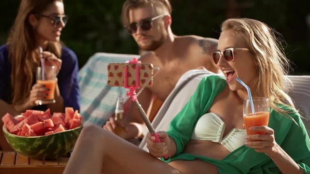 Two gorgeous females in sunglasses smiling talking with bearded topless male with piercing making selfie drinking cocktails. In slowmotion