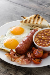 Traditional full English breakfast with fried eggs, sausages, beans, mushrooms, grilled tomatoes and bacon on wooden background
