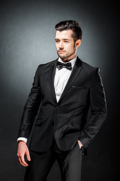 portrait of confident handsome man in black suit with bowtie posing in dark studio background
