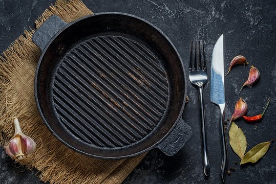 Black iron empty grill pan, seasonings, fork and knife on cement black texture background