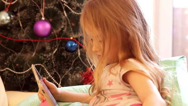 Girl playing with tablet PC near Christmas tree
