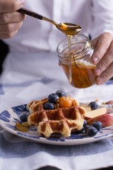 Blueberry apple waffle with honey set on a dish with a chef's hand holding spoon for a jar of honey ready to serve.