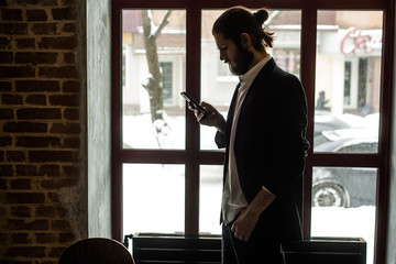 young bearded man in a jacket and white shirt looks into the smartphone against the window
