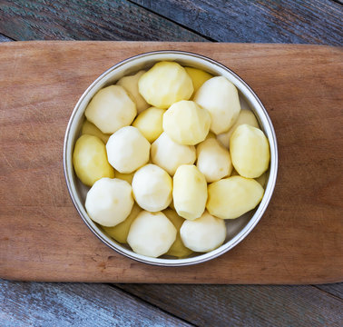 raw peeled potatoes in a circular bowl on an old cutting board on rustic wooden table closeup, top view