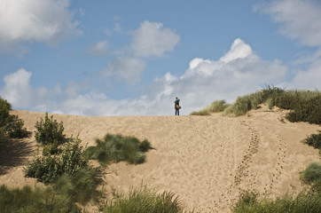 Fototapeta premium man standing looks off into the distance after running to the top of dune in desert with bushes plants, blue sky, summer with white clouds