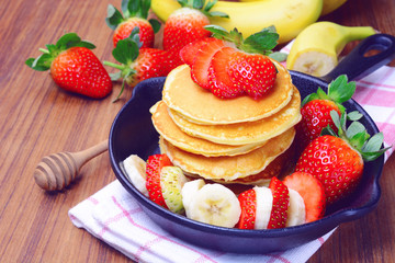 Delicious pancake with sliced of strawberry and banana on black iron pan and fresh fruit on wooden table.