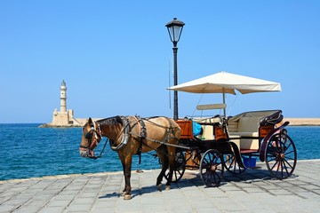 Horse drawn carriage on the quayside with the Venetian lighthouse at the harbour entrance to the rear, Chania, Crete.