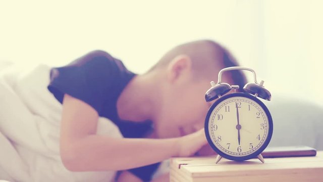 Vintage color tone selective focus at vintage alarm clock show at six o clock and out focus young asian boy sleeping and wake up to play smart phone on bed.