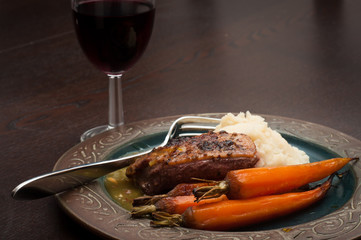 Roasted duck breast in orange sauce with carrots and celeriac mash. Served on a dark wooden table with a glass of red wine.