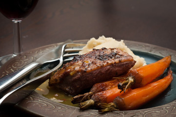 Roasted duck breast in orange sauce with carrots and celeriac mash. Served on a dark wooden table with a glass of red wine.