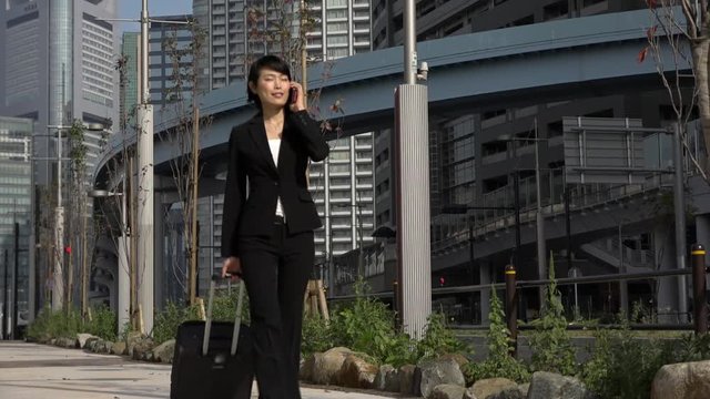 Happy beautiful Japanese female manager speaking on cell phone. Asian businesswoman, confident girl, busy woman talking with mobile telephone and walking with luggage during business trip. Slow motion