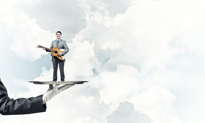 Businessman on metal tray playing acoustic guitar against blue sky background