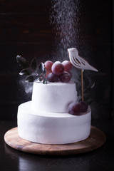 White two-tiered cake decorated with a bird grapes and leaves on a dark wooden background with dark light