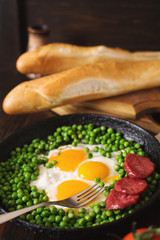 Fried eggs with sausage and green peas on a black frying pan on a wooden background