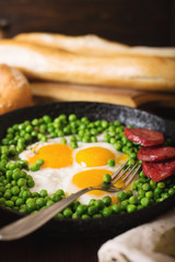 Fried eggs with sausage and green peas on a black frying pan on a wooden background