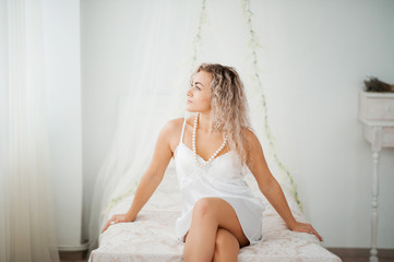 Curly blonde girl in a white silk gown boudoir robe on bed