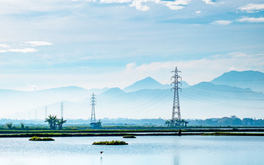Transmission tower, electricity pylon or a power pylon, standing majestically lined up to the horizon, in front of large lake or pond, also with beautiful cloud and mountain in the background, capture