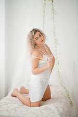 Curly blonde girl in a white silk gown boudoir robe on bed.