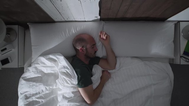 Young adult man can't sleep at night because of an irritating mosquito. He shakes his hand trying to get away the mosquito. Finally, he hides under the sheets.