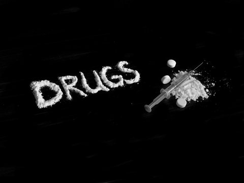 Cocaine drug powder in drugs word shaped, injection syringe on cocaine powder pile and pills on black background in black and white colors