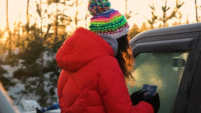 Young woman in bright red jacket and bright hat remove snow and ice from the car with snow brush under the sunlight