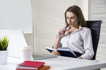 Close up of a serious and beautiful businesswoman wearing a white blouse and sitting at her desk with a notebook and glasses. Mock up
