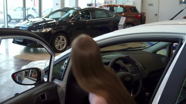 Pretty young woman sitting into the car at the dealership. Close up of attractive caucasian girl opening car door. Beautiful brown haired lady putting her hands on the steering wheel