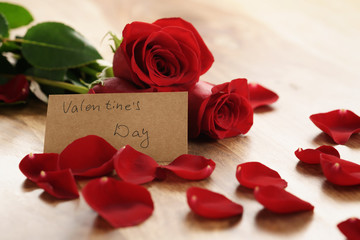 warm photo of three red roses with petals on wood table and paper card for valentines day, shallow focus