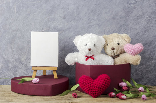 Love concept of teddy bear in red heart gift box on wood table and blank canvas frame on easel painting for valentines day and wedding