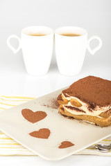 Tiramisu /
Tiramisu cake in a plate with a cocoa decoration and two cups of coffee on a white background