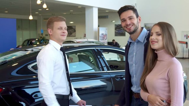 Blond salesman giving the car key to the couple at the dealership. Side view of sales manager and customers having deal at the car showroom. Attractive young people smiling against background of