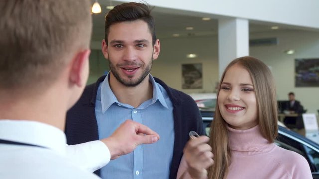 Attractive caucasian couple buying the car at the dealership. Pretty brown haired girl taking car key from the hand of salesman. Blond sales manager standing backwards against background of brunette