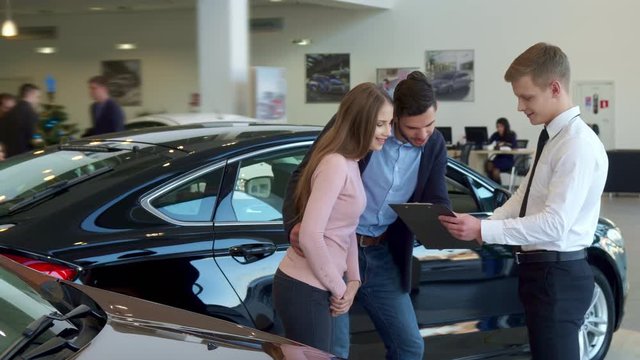 Long shot of young couple looking at the contract at the dealership. Blond caucasian salesman holding clipboard in his hand. Male sales manager and his clients standing against background of black car
