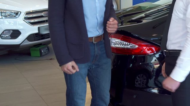 Customer getting car key at the dealership. Close up of male hands shaking against background of new cars. Two men standing near black sedan at the car showroom