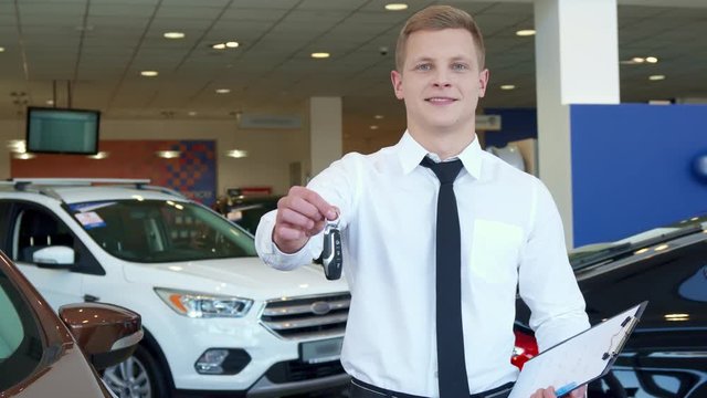 Attractive sales manager showing car key at the dealership. Young caucasian salesman writing on his clipboard. Handsome blond man in white shirt and black tie smiling for the camera