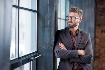 Portrait of caucasian bearded businessman dressed in suit with eyeglasses near window in the loft interior office