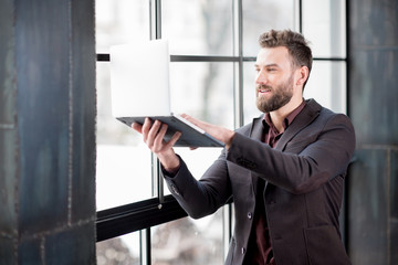 Handsome bearded businessman dressed in the suit trying to get internet connection with laptop near the big window in the loft interior studio