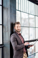 Handsome bearded businessman dressed in the suit standing with laptop near the big window in the loft interior studio