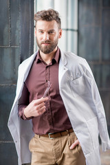 Portrait of handsome confident dentist with medical gown and dental tools standing near the window in the modern dark interior clinic or office