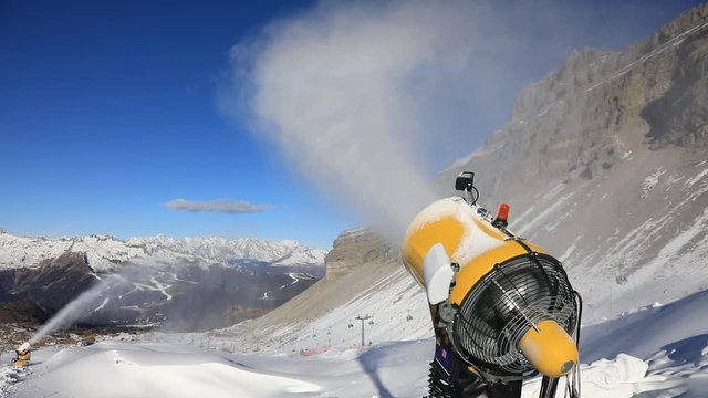Yellow snow cannon stands on a snowy mountain in the winter and works by producing a column of snow on the background of beautiful Alpine mountains and the ski slopes
