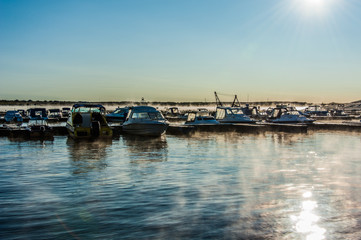 Fototapeta na wymiar Boats and yachts moored at the port. Glittering sea surface against sunrise. Morning fog.