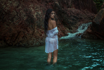 Romantic brunette beauty wearing white wet shirt standing in the water between rocks near beautiful...